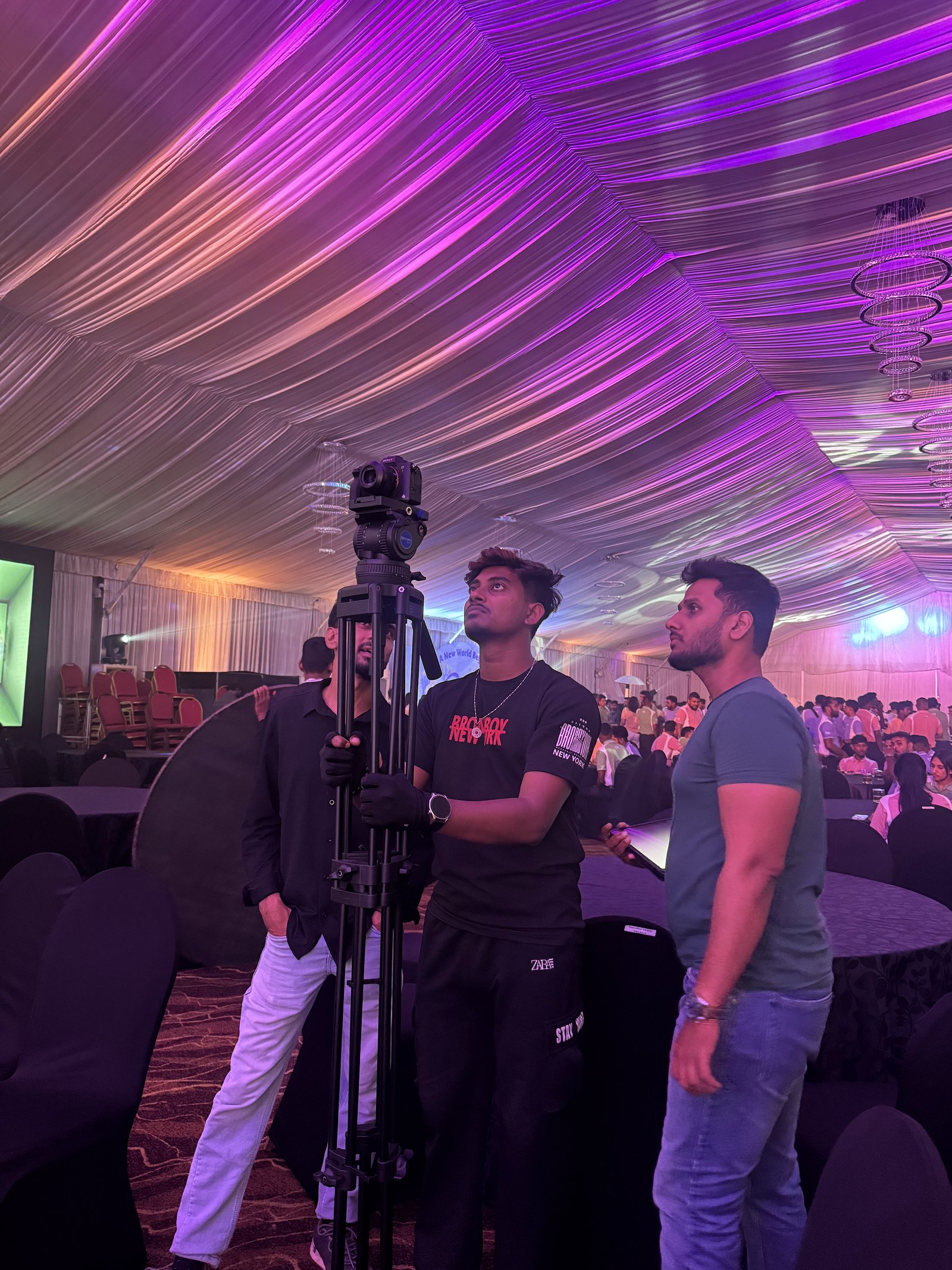 Camera crew with a tripod-mounted camera among seated guests under a draped event ceiling